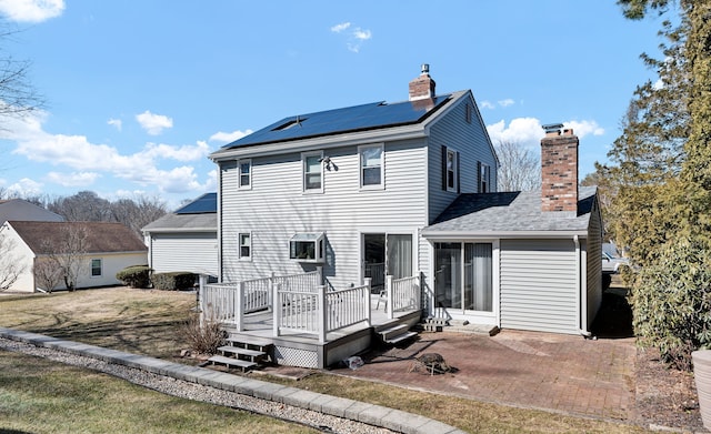 back of property with a chimney, a shingled roof, roof mounted solar panels, a patio area, and a deck