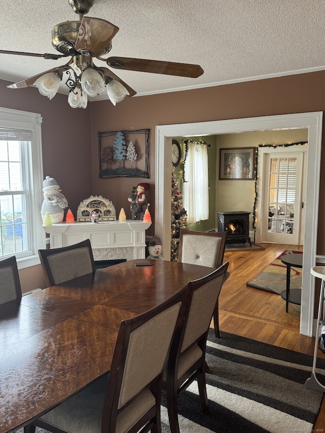 dining area featuring a textured ceiling, a warm lit fireplace, wood finished floors, ornamental molding, and a wood stove