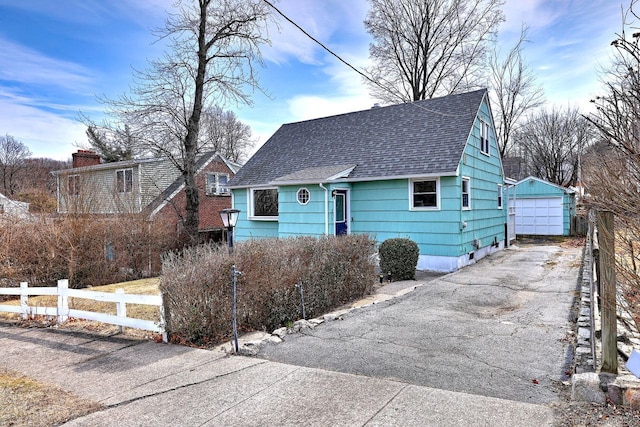 view of front of house with driveway, a detached garage, roof with shingles, an outbuilding, and fence