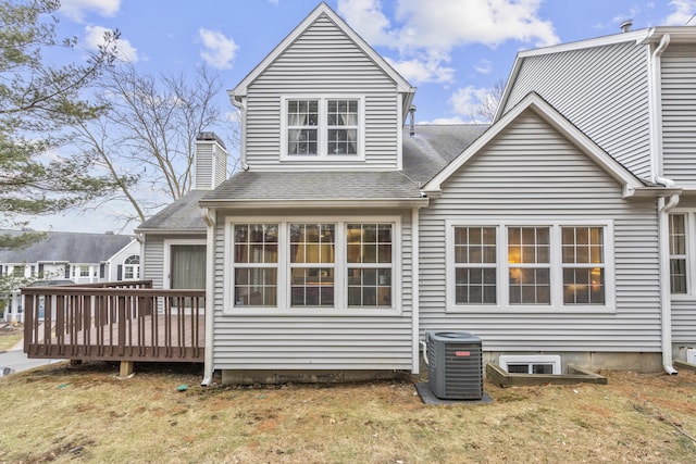 rear view of property featuring central AC unit, a shingled roof, a lawn, a wooden deck, and a chimney