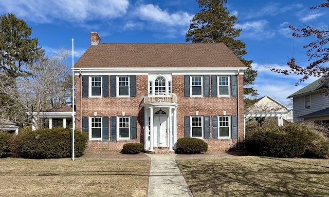 colonial home featuring a front yard, brick siding, roof with shingles, and a chimney