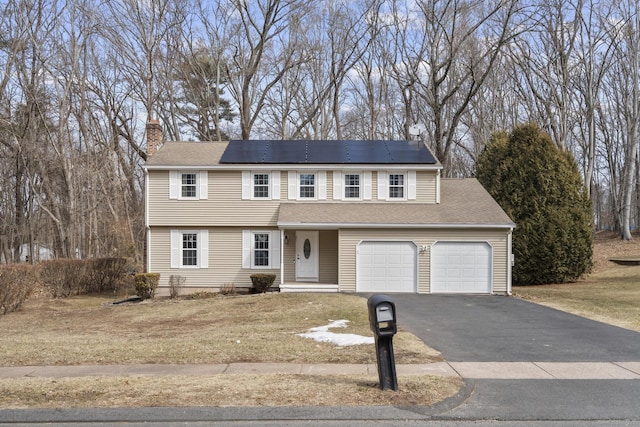 colonial inspired home with aphalt driveway, an attached garage, solar panels, a chimney, and a front yard