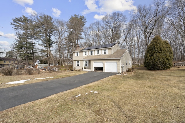 colonial inspired home featuring an attached garage, driveway, roof mounted solar panels, a front lawn, and a chimney