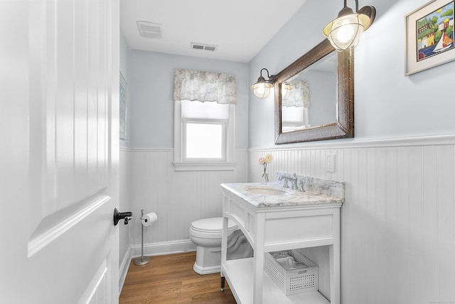 half bathroom with visible vents, toilet, wainscoting, vanity, and wood finished floors