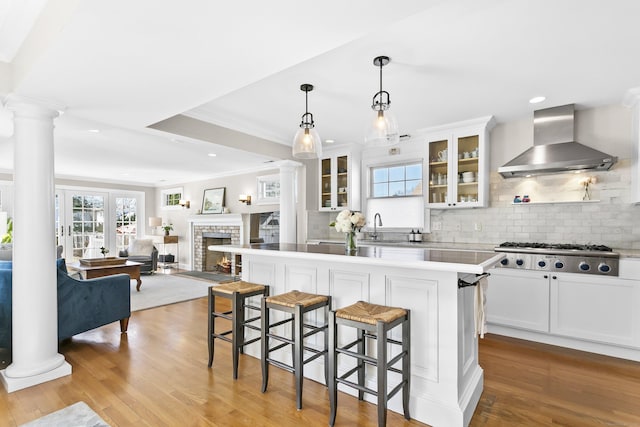 kitchen featuring a breakfast bar, stainless steel gas stovetop, wall chimney exhaust hood, light wood finished floors, and ornate columns