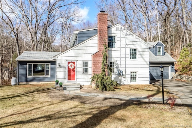 rear view of house with driveway, a yard, a shingled roof, a garage, and a chimney