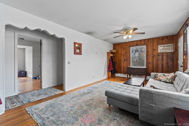 living area featuring wooden walls, baseboards, ceiling fan, wood finished floors, and an AC wall unit