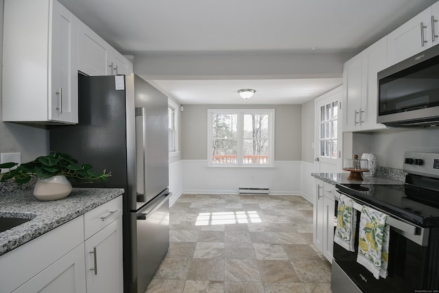 kitchen featuring white cabinetry, appliances with stainless steel finishes, baseboard heating, wainscoting, and light stone countertops