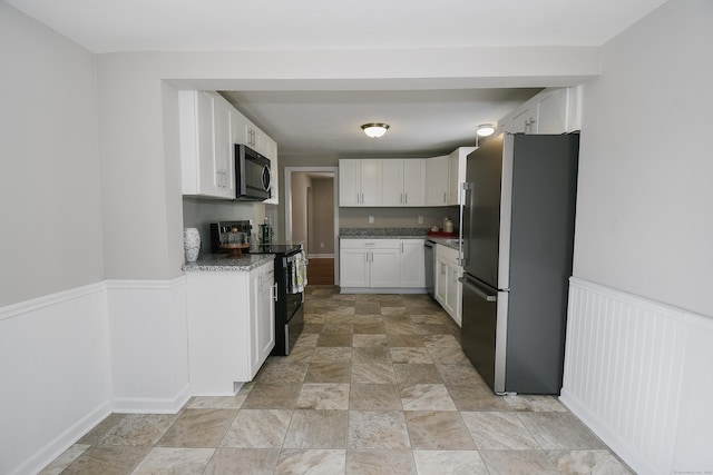 kitchen with appliances with stainless steel finishes, stone finish floor, a wainscoted wall, and white cabinetry