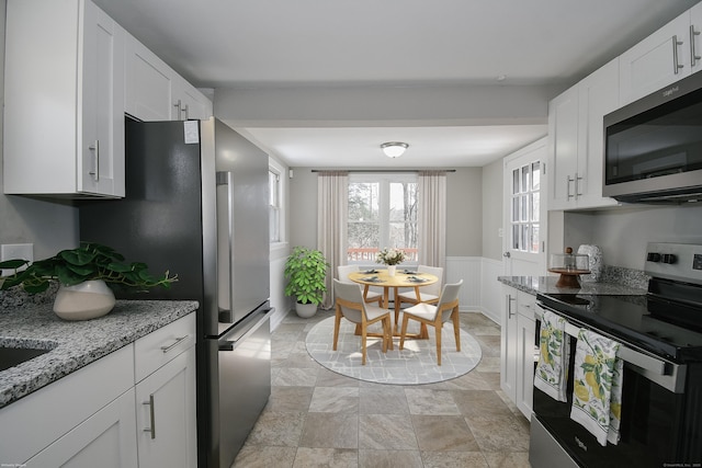 kitchen featuring stainless steel appliances, wainscoting, white cabinetry, and light stone countertops