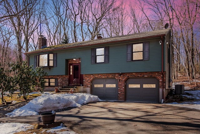 raised ranch featuring a garage, central AC, brick siding, driveway, and a chimney