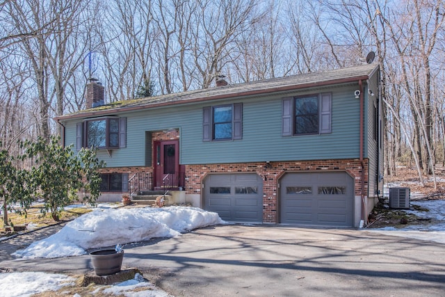 bi-level home featuring brick siding, a chimney, central air condition unit, a garage, and driveway