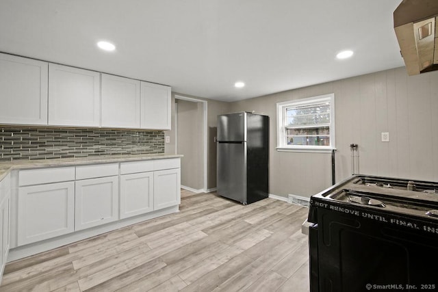 kitchen with black gas range oven, light wood-style flooring, freestanding refrigerator, white cabinetry, and backsplash