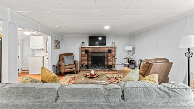 living room with a brick fireplace, ornamental molding, wood finished floors, baseboard heating, and stacked washing maching and dryer