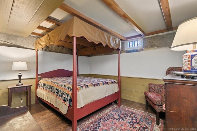 bedroom with beamed ceiling, wood finished floors, and wainscoting