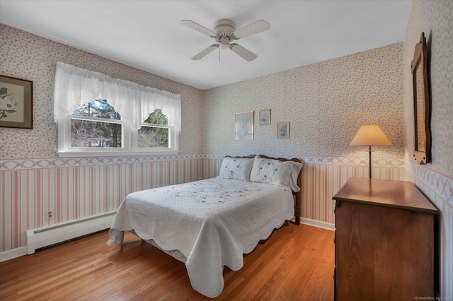 bedroom featuring a wainscoted wall, light wood-style flooring, baseboard heating, and wallpapered walls