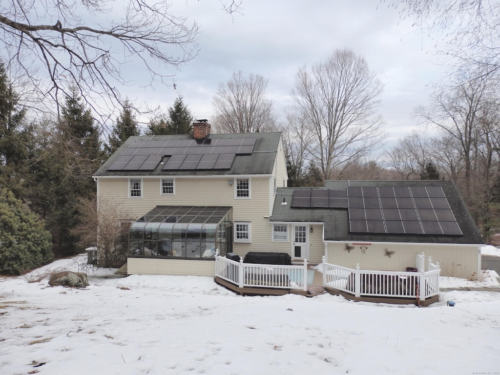 snow covered house with a chimney, a wooden deck, a sunroom, and roof mounted solar panels