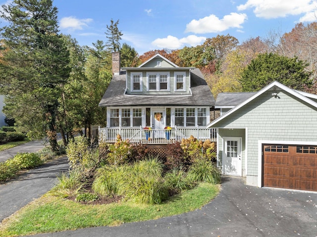 shingle-style home with an attached garage, covered porch, a chimney, and aphalt driveway