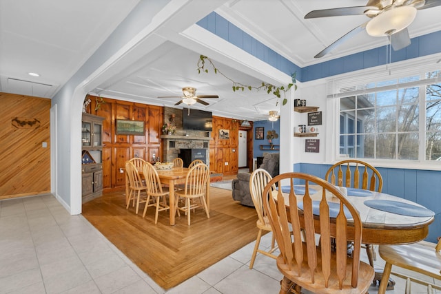 dining room featuring a large fireplace, a ceiling fan, ornamental molding, wood walls, and light tile patterned flooring