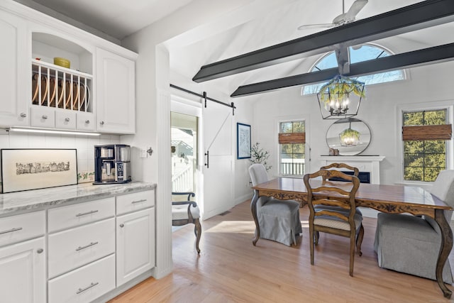 dining area featuring light wood finished floors, lofted ceiling with beams, a fireplace, a barn door, and ceiling fan with notable chandelier