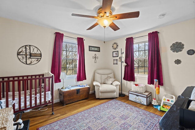 bedroom featuring a nursery area, ceiling fan, baseboards, and wood finished floors