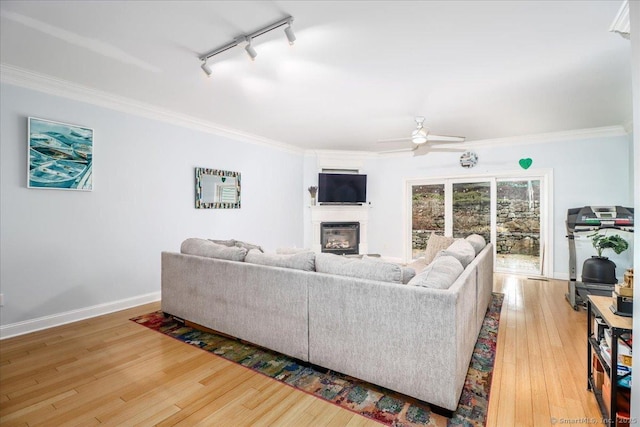 living area featuring light wood-style flooring, a glass covered fireplace, crown molding, baseboards, and ceiling fan