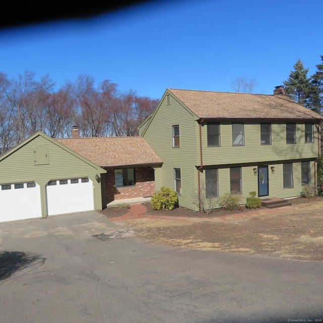 colonial-style house with brick siding, an attached garage, roof with shingles, a chimney, and driveway