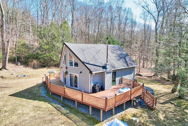 rear view of property featuring a forest view, a lawn, a wooden deck, and roof with shingles