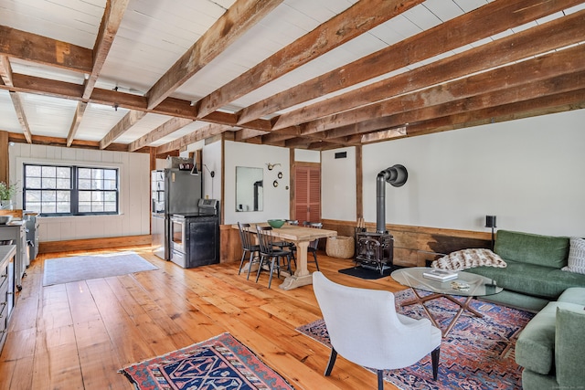 living room with light wood finished floors, beamed ceiling, wood walls, wainscoting, and a wood stove