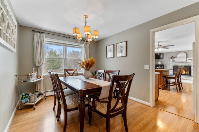 dining space featuring baseboards, a stone fireplace, light wood-style floors, a baseboard heating unit, and ceiling fan with notable chandelier