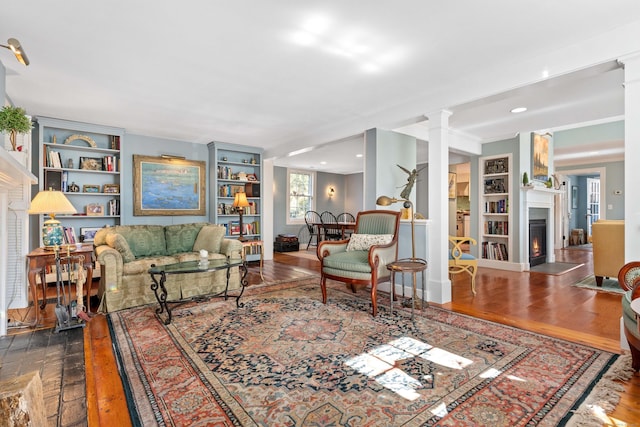 living area featuring a fireplace with flush hearth, built in features, ornate columns, and wood finished floors