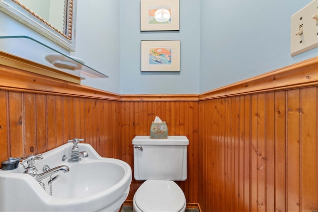 bathroom featuring wooden walls, wainscoting, toilet, and a sink