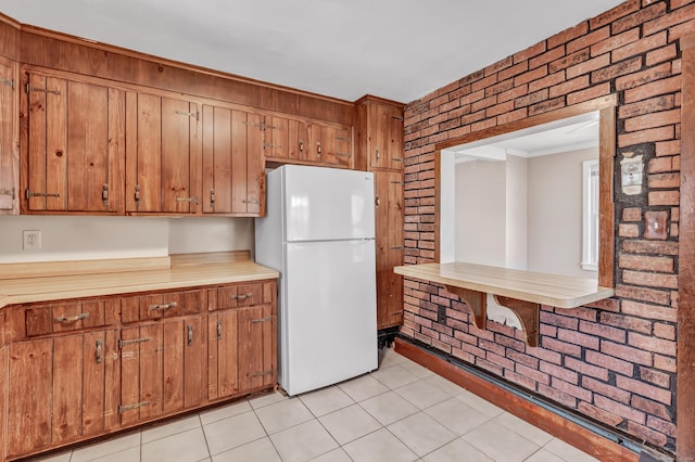 kitchen with light tile patterned floors, brown cabinetry, brick wall, freestanding refrigerator, and light countertops