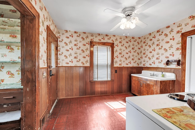 bathroom with a sink, a ceiling fan, wainscoting, and wallpapered walls