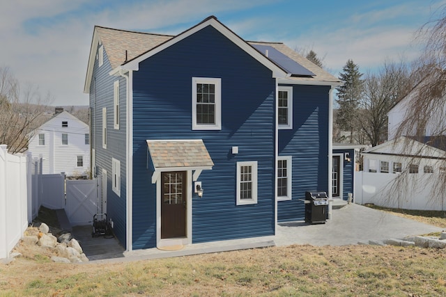 back of house with a gate, roof mounted solar panels, fence, roof with shingles, and a patio area