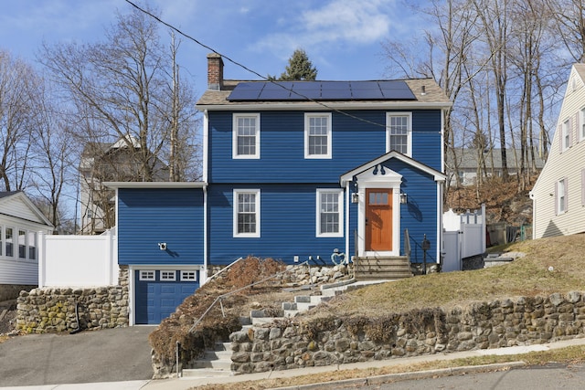 colonial home featuring fence, a garage, roof mounted solar panels, and a chimney