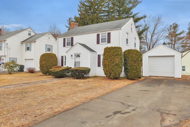 colonial house featuring a shingled roof, a detached garage, aphalt driveway, a chimney, and an outbuilding