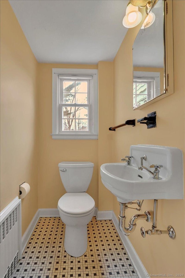 half bath with tile patterned flooring, baseboards, toilet, radiator heating unit, and a sink