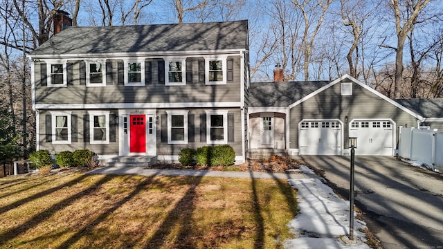 colonial house with driveway, a chimney, roof with shingles, an attached garage, and fence