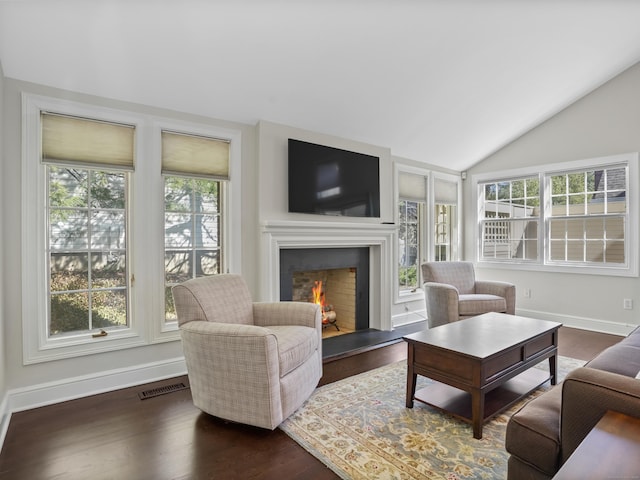 living area featuring baseboards, visible vents, dark wood-style flooring, a lit fireplace, and vaulted ceiling