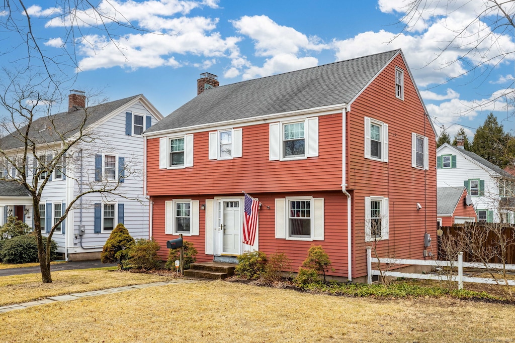 colonial inspired home featuring a shingled roof, a chimney, fence, and a front lawn