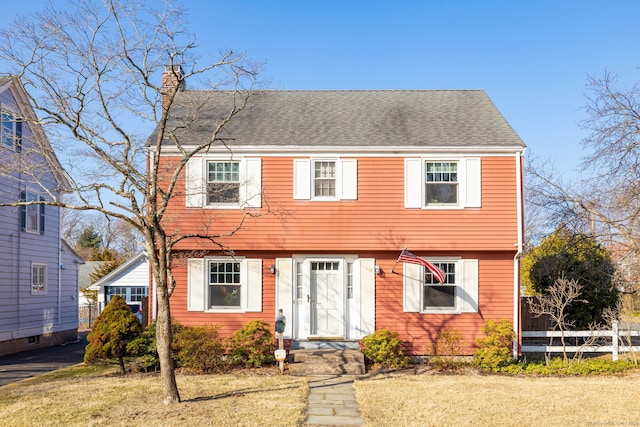 colonial-style house with a front lawn, fence, roof with shingles, and a chimney