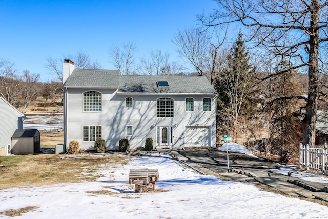 snow covered property with an outbuilding, a shed, a chimney, concrete driveway, and a garage