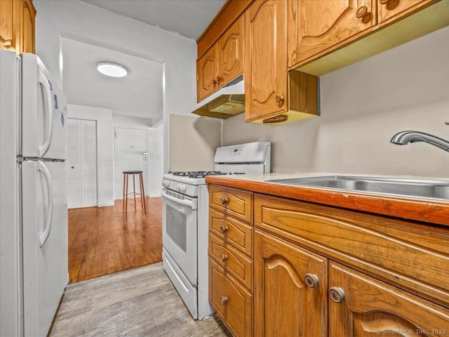 kitchen with white appliances, light wood finished floors, a sink, under cabinet range hood, and brown cabinets