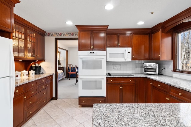 kitchen with backsplash, glass insert cabinets, light stone counters, light tile patterned floors, and white appliances