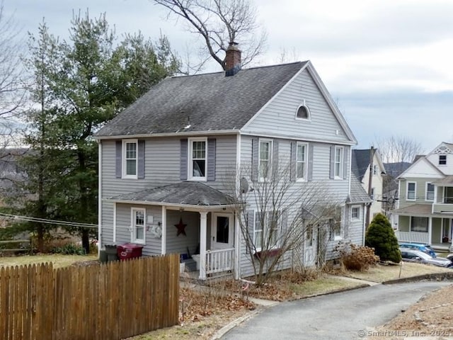 traditional-style house featuring a porch, a shingled roof, a chimney, and fence