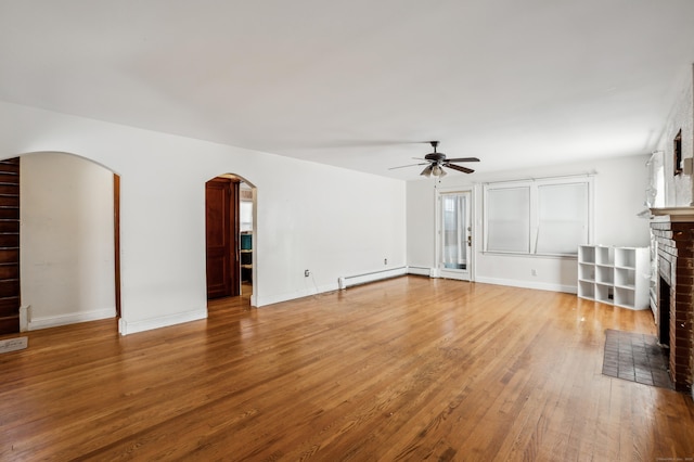 unfurnished living room featuring a ceiling fan, light wood-style flooring, arched walkways, a baseboard heating unit, and a brick fireplace