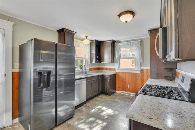 kitchen featuring a sink, stainless steel appliances, dark brown cabinetry, wood walls, and wainscoting