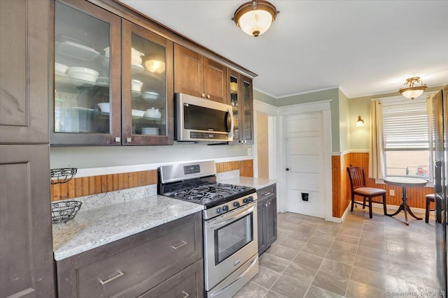 kitchen with a wainscoted wall, stainless steel appliances, wood walls, glass insert cabinets, and light stone countertops