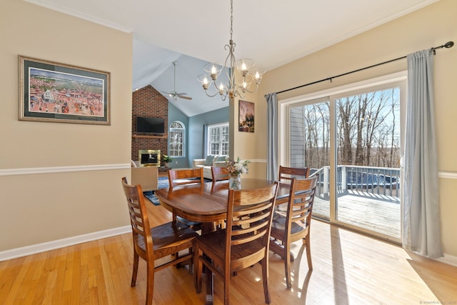 dining space with baseboards, a fireplace, vaulted ceiling, light wood-style floors, and ceiling fan with notable chandelier